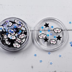 May include: A clear plastic container filled with small, white cloud-shaped charms with black outlines and pink cheeks. The container is surrounded by blue and silver glitter stars.