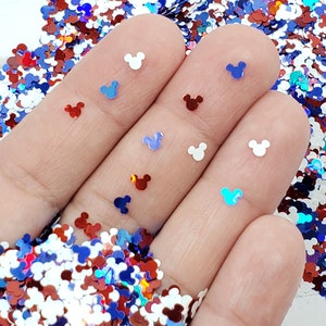 Pu&ograve; includere: Una mano che tiene una variet&agrave; di glitter iridescente rosso, bianco e blu a forma di teste di Topolino.