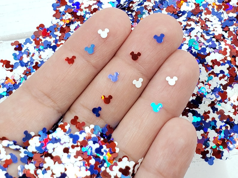 Pu&ograve; includere: Una mano che tiene una variet&agrave; di glitter iridescente rosso, bianco e blu a forma di teste di Topolino.