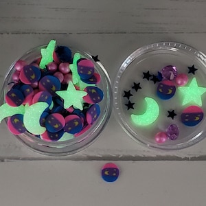 May include: Two clear containers filled with glow-in-the-dark sprinkles. The sprinkles are shaped like stars, moons, and crescent moons. The sprinkles are pink, blue, and green.