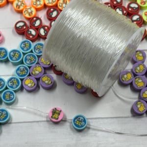 Op de afbeelding: Een verzameling kleurrijke ronde kralen met cartoonkarakterontwerpen, waaronder oranje, rood, roze, blauw en paars. Een spoel met doorzichtige draad ligt naast de kralen, waarvan sommige op de draad zijn geregen. De kralen zijn waarschijnlijk voor het maken van sieraden.