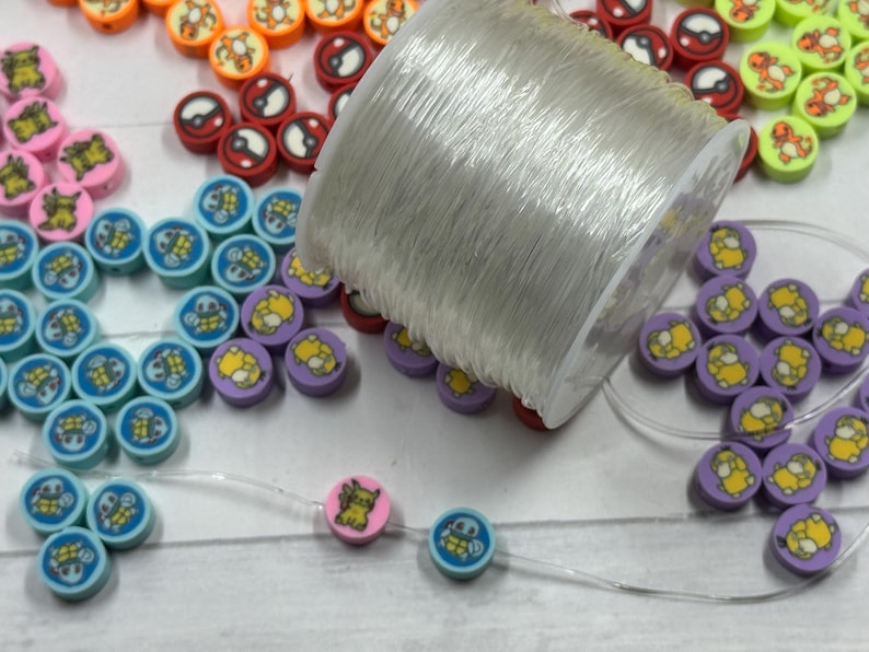 Op de afbeelding: Een verzameling kleurrijke ronde kralen met cartoonkarakterontwerpen, waaronder oranje, rood, roze, blauw en paars. Een spoel met doorzichtige draad ligt naast de kralen, waarvan sommige op de draad zijn geregen. De kralen zijn waarschijnlijk voor het maken van sieraden.