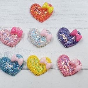 May include: Seven heart-shaped resin embellishments with glitter and a pink bow on each. The hearts are in various colors: pink, orange, purple, blue, yellow, and white.