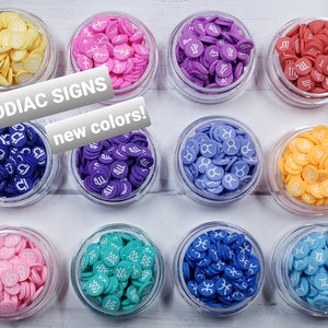 May include: Twelve small jars filled with colorful round resin zodiac signs. Each jar contains a different zodiac sign and color. The jars are arranged in a grid pattern on a white wooden surface. The text "Zodiac Signs new colors!" is written in white letters on a black background.