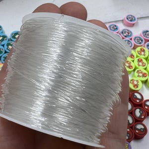 Op de afbeelding: Een spoel met doorzichtig elastisch draad wordt in een hand gehouden, met diverse kleurrijke kralen eromheen. De kralen tonen cartoonkarakters en ontwerpen, waaronder kikkers, tijgers en Pokeball-graphics. De kralen zijn in tinten blauw, roze, rood en geel.