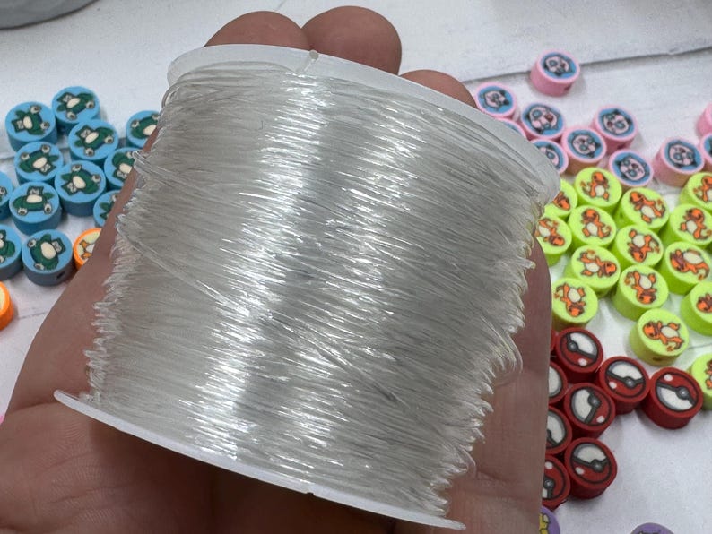 Op de afbeelding: Een spoel met doorzichtig elastisch draad wordt in een hand gehouden, met diverse kleurrijke kralen eromheen. De kralen tonen cartoonkarakters en ontwerpen, waaronder kikkers, tijgers en Pokeball-graphics. De kralen zijn in tinten blauw, roze, rood en geel.