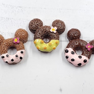 May include: Three chocolate donuts with different colored icings and sprinkles. The donuts are shaped like Mickey Mouse ears.