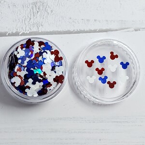 Pu&ograve; includere: Un contenitore di plastica trasparente pieno di glitter rosso, bianco e blu a forma di teste di Topolino. Il contenitore ha un coperchio con alcune teste di Topolino sopra.