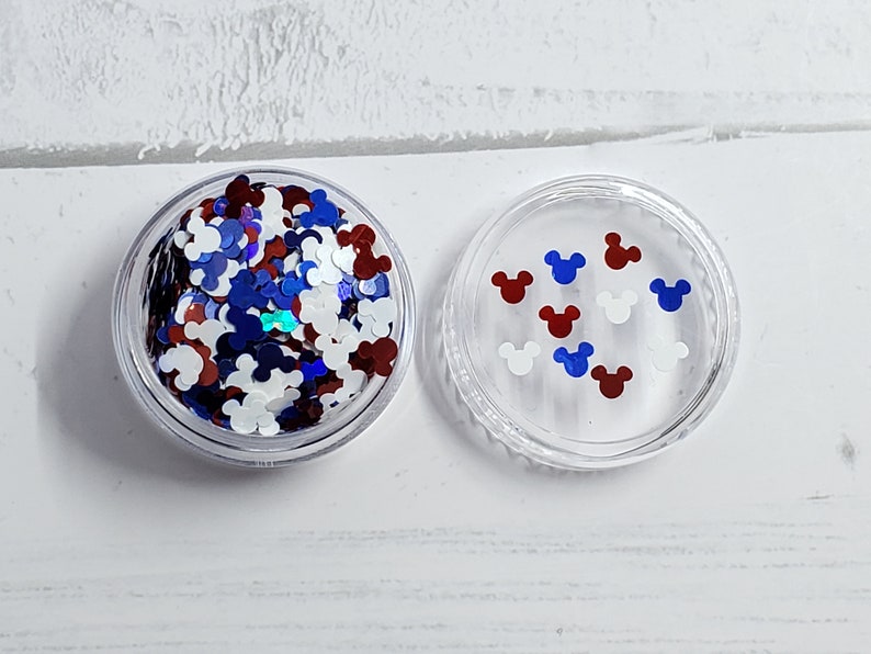 Pu&ograve; includere: Un contenitore di plastica trasparente pieno di glitter rosso, bianco e blu a forma di teste di Topolino. Il contenitore ha un coperchio con alcune teste di Topolino sopra.