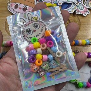May include: A clear, iridescent bag filled with colorful beads in various shapes and sizes. The bag contains pink, purple, blue, yellow, and orange beads, along with small wooden clothespins and decorative charms. The bag has a cartoon bunny illustration.