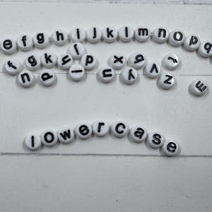 May include: White, round alphabet beads with black letters are scattered across a white surface. The word "lowercase" is spelled out with the beads. Other beads are arranged in a curved line, displaying various letters.