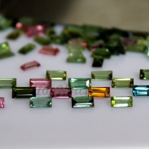 May include: Assortment of faceted, rectangular gemstones in various colors, including green, pink, blue, and orange. The gemstones are arranged in a row on a white surface.