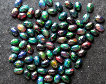 Puede incluir: Una colección de ópalos negros ovalados con colores iridiscentes verdes, azules y rojos. Los ópalos están dispuestos en un montón suelto sobre una superficie gris oscura.