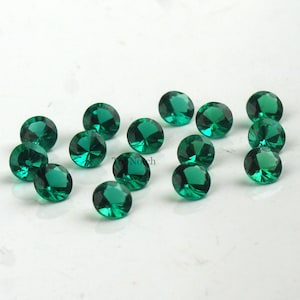 May include: A collection of 15 round, faceted, emerald green gemstones. The gemstones are scattered on a white surface.