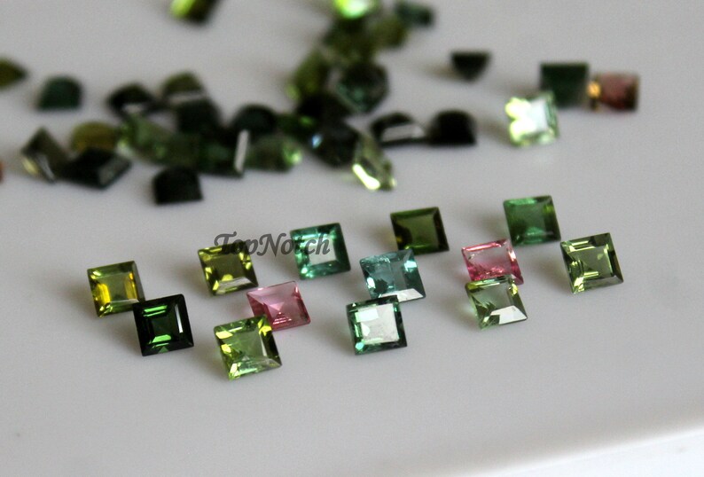May include: Assortment of green, pink, and blue square-shaped gemstones. The gemstones are faceted and have a shiny finish.