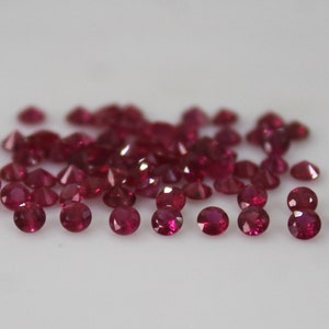 May include: A scattering of small, round, faceted rubies on a white surface. The gemstones are a deep red colour, with facets that catch the light, creating a sparkling effect. The gems vary slightly in size.