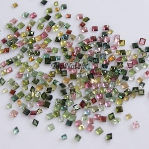 May include: A collection of loose, faceted, square-shaped gemstones in various shades of green, pink, and brown. The gemstones are scattered on a white surface.