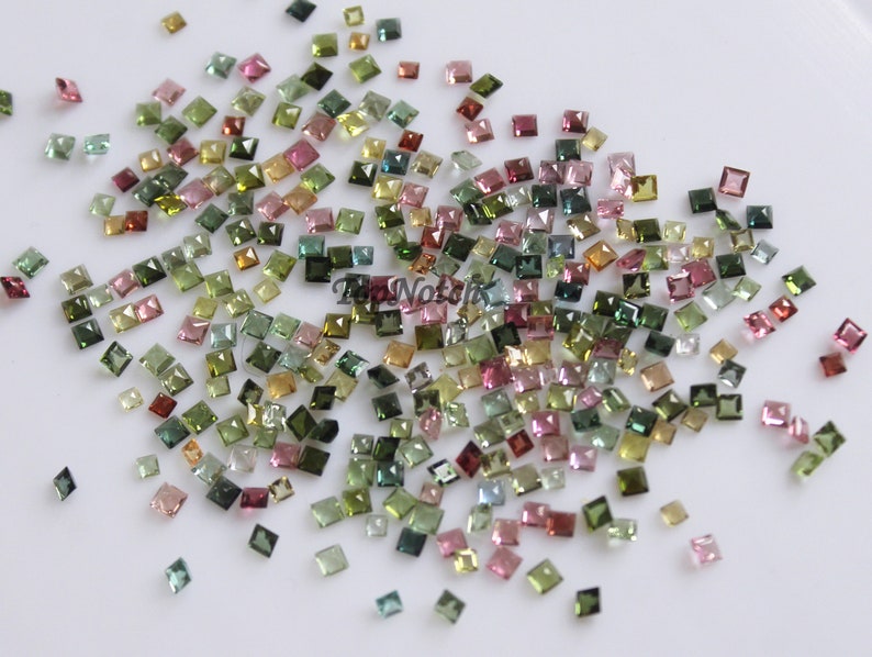 May include: A collection of loose, faceted, square-shaped gemstones in various shades of green, pink, and brown. The gemstones are scattered on a white surface.