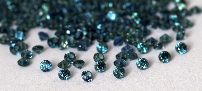Op de afbeelding: Een stapel teal blauwe edelstenen, waarvan sommige verspreid liggen op een wit oppervlak.