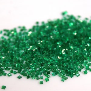 May include: A pile of small, square, emerald green gemstones. The gemstones are scattered on a white surface.
