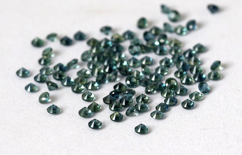 Op de afbeelding: Een strooiing van teal blauwe edelstenen, waarschijnlijk saffieren, op een wit oppervlak. De edelstenen zijn gefacetteerd en hebben verschillende groottes.