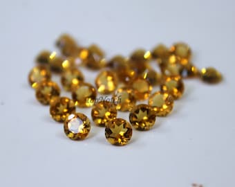 May include: A group of 20 round, faceted, golden yellow gemstones. The gemstones are scattered on a white surface.