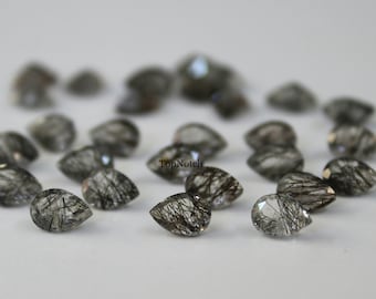 May include: A collection of pear-shaped, clear quartz gemstones with black inclusions. The stones are arranged in a circle on a white surface.
