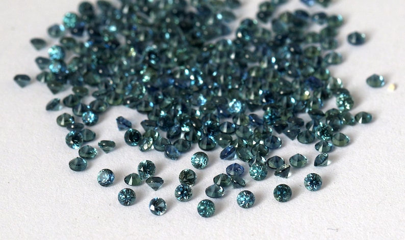 Op de afbeelding: Een hoop losse teal blauwe edelstenen, verspreid over een wit oppervlak.