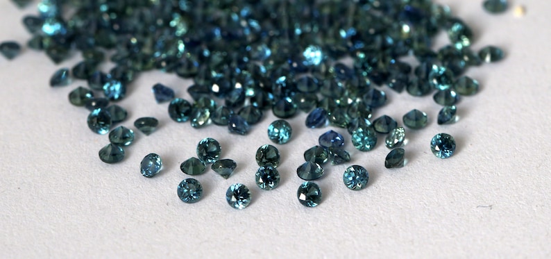 Op de afbeelding: Een hoop losse teal blauwe edelstenen, sommige zijn verspreid over een wit oppervlak.