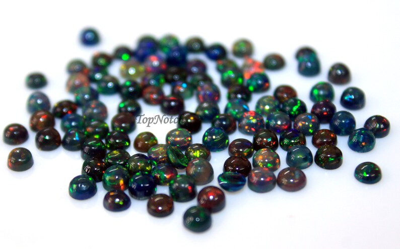 May include: A collection of round, iridescent black opal cabochons. The opals have a variety of colors, including green, blue, red, and orange. The opals are scattered on a white surface.