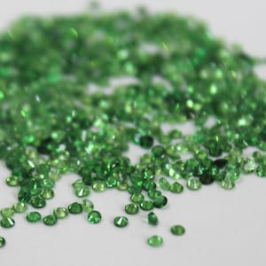 May include: A large pile of small, round, green gemstones. The gemstones are scattered on a white surface.