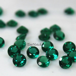 May include: A group of 20 round, emerald green gemstones. The gemstones are scattered on a white surface.