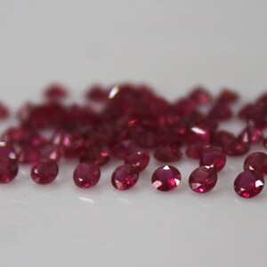 May include: A collection of small, oval-cut rubies. The gemstones are a deep, rich red color and are scattered across a white surface. The rubies are faceted, catching the light and creating a sparkling effect.