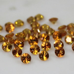 Könnte beinhalten: Eine Gruppe von 30 runden, facettierten, orangefarbenen Edelsteinen. Die Edelsteine sind auf einer weißen Oberfläche verstreut.