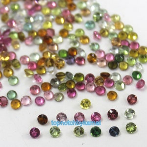 Op de afbeelding: Assortiment van ronde, gefacetteerde edelstenen in verschillende kleuren, waaronder roze, groen, geel en blauw. De edelstenen zijn verspreid over een wit oppervlak.  topnotchbykamal