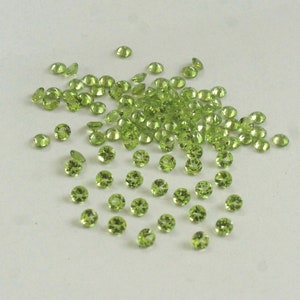 May include: A large pile of small, round, light green peridot gemstones. The gemstones are scattered on a white surface.