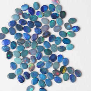 May include: A collection of oval-shaped opals in various shades of blue, teal, and green. The opals display iridescent flashes of color, including hints of red, yellow, and purple. The gemstones are scattered on a white surface, showcasing their natural beauty.