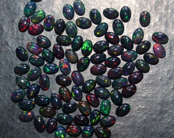 Puede incluir: Una colección de ópalos negros de forma ovalada con destellos iridiscentes verdes, azules y rojos. Los ópalos están dispuestos en un patrón suelto en forma de corazón sobre una superficie gris oscura.