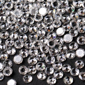 May include: A collection of small, clear, faceted gemstones scattered on a dark surface. The gemstones have a rose-cut design, reflecting light and creating a sparkling effect. The overall impression is one of luxury and elegance.