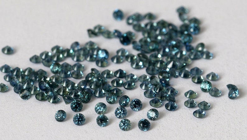 Op de afbeelding: Een verzameling losse teal blauwe edelstenen, waarschijnlijk saffieren, verspreid over een wit oppervlak.