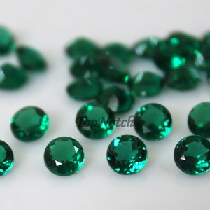 May include: A collection of round, faceted emerald green gemstones. The gems are scattered across a white surface, with some in focus and others slightly blurred. The gems have a brilliant cut, reflecting light.