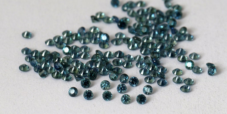 Op de afbeelding: Een losse stapel teal blauwe edelstenen, waarschijnlijk saffieren, op een wit oppervlak.