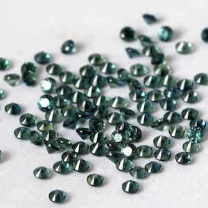 Op de afbeelding: Een verzameling losse teal blauwe edelstenen, waarschijnlijk saffieren, verspreid over een wit oppervlak. De edelstenen zijn gefacetteerd en hebben een briljante glans.