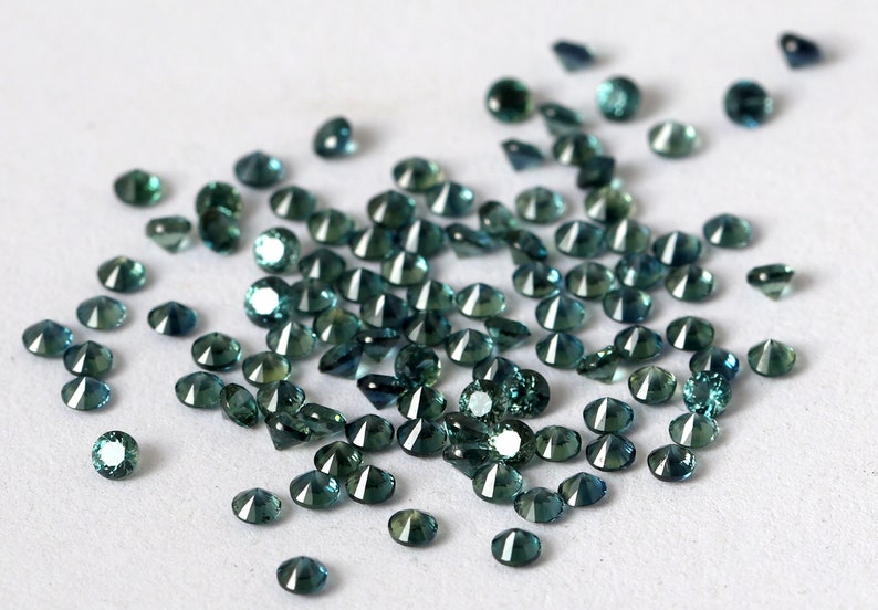 Op de afbeelding: Een verzameling losse teal blauwe edelstenen, waarschijnlijk saffieren, verspreid over een wit oppervlak. De edelstenen zijn gefacetteerd en hebben een briljante glans.