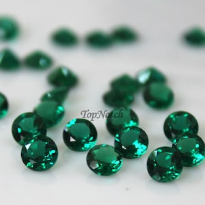 May include: A collection of round, faceted, emerald green gemstones. The gemstones are scattered on a white surface.