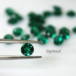 May include: A close-up of a single, round, green gemstone held in tweezers. The gemstone is surrounded by a scattering of similar gemstones on a white surface. The text "TopNotch" is visible in the lower right corner of the image.