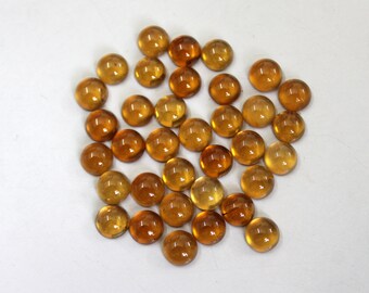 May include: A collection of small, round, amber-colored glass beads. The beads have a smooth, polished surface and vary slightly in shade, from light yellow to deep orange. They are scattered on a white surface, creating a warm, inviting aesthetic.
