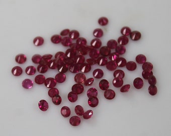 May include: A collection of loose, round, red gemstones. The gemstones are scattered on a white surface.