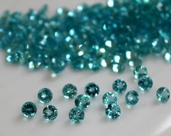 May include: A collection of small, round, turquoise gemstones. The gemstones are scattered on a white surface.