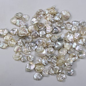 Puede incluir: Una gran colección de perlas de agua dulce blancas e iridiscentes de forma irregular. Las perlas están esparcidas sobre una superficie blanca.